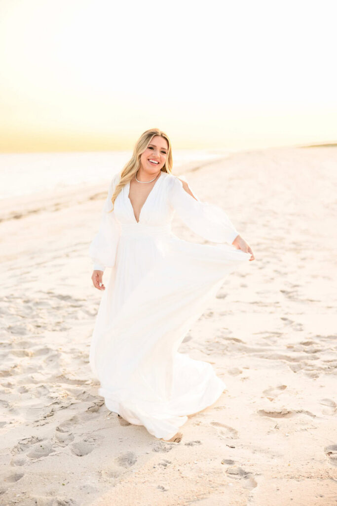 Bride walking along a soft sandy beach at sunset, holding her dress with a joyful expression in a light and airy engagement-style portrait