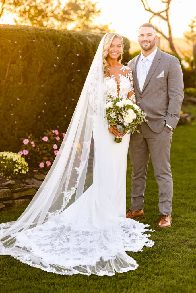 Bride and groom standing together in a garden at golden hour, holding a bouquet and smiling in a timeless wedding portrait on Long Island
