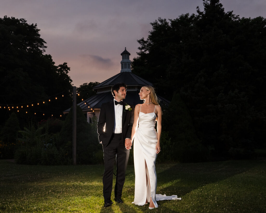 Evening wedding portrait by a Luxury Wedding Photographer Long Island of a couple walking near a garden pavilion under soft string lights
