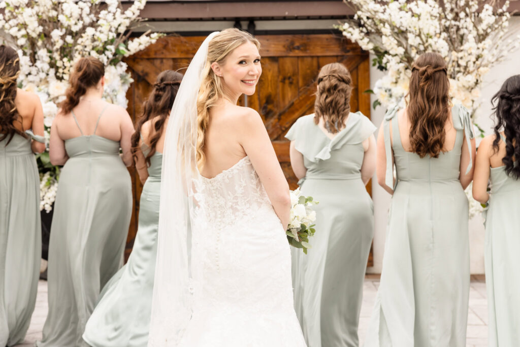 Bride smiling over her shoulder while standing with bridesmaids in soft green dresses in front of a floral wedding backdrop