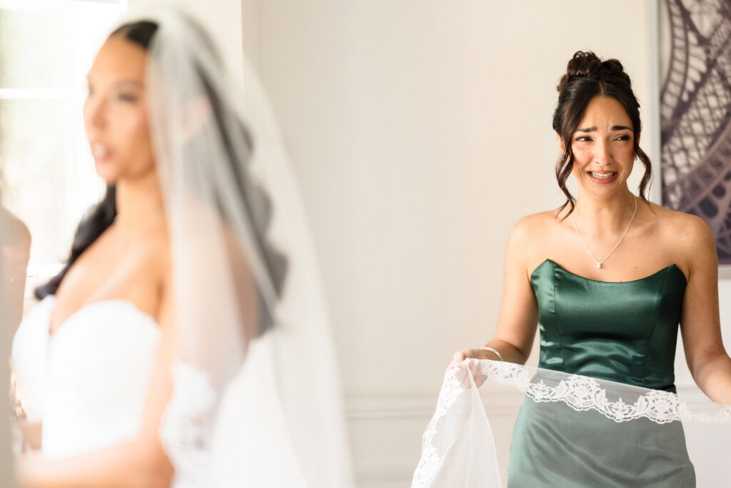 Bridesmaid holding a veil and reacting emotionally while seeing the bride during wedding morning preparations in soft natural light