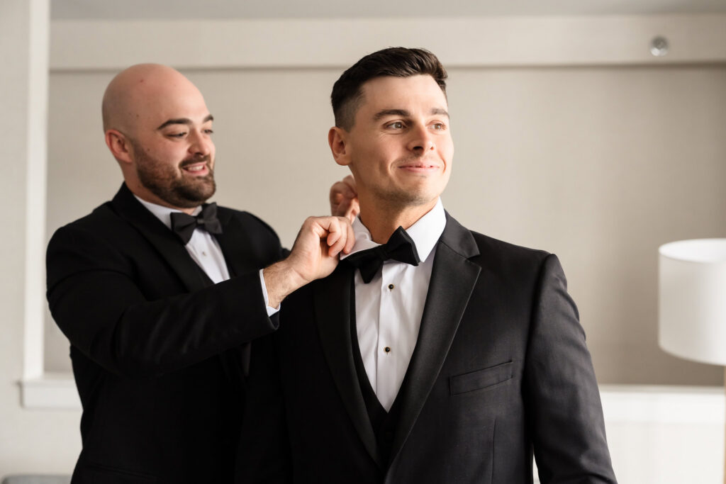 Groom smiling while his groomsman adjusts his bowtie during wedding day preparations in soft natural light
