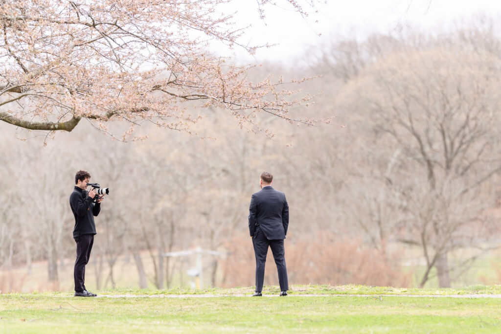 Second wedding photographer capturing groom portraits outdoors beneath spring trees before the ceremony.