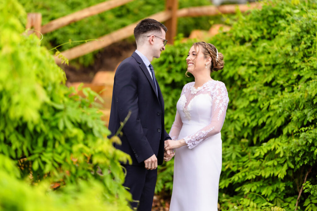 Bride and groom holding hands and smiling together in a lush green garden on their wedding day.