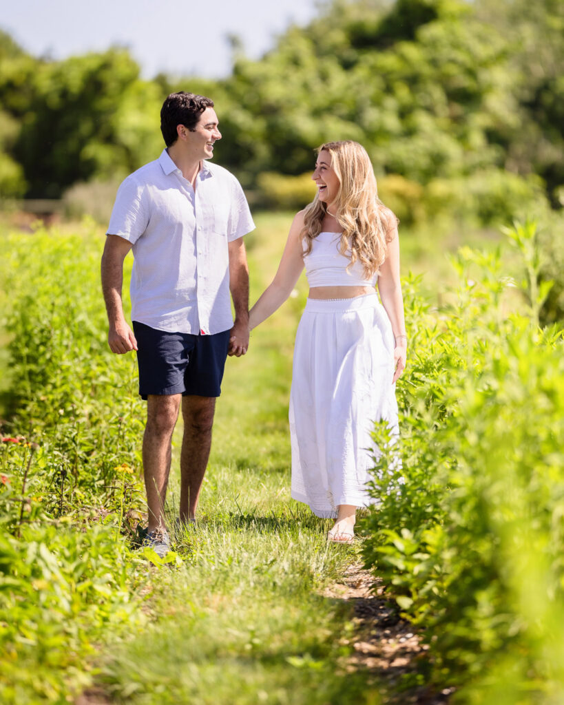 Engaged couple walking hand in hand through a sunny green field during their summer engagement session.