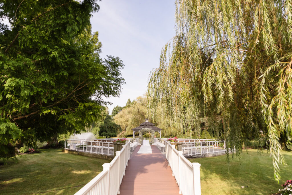 Outdoor garden wedding ceremony location with a white bridge leading to a gazebo surrounded by trees and water.