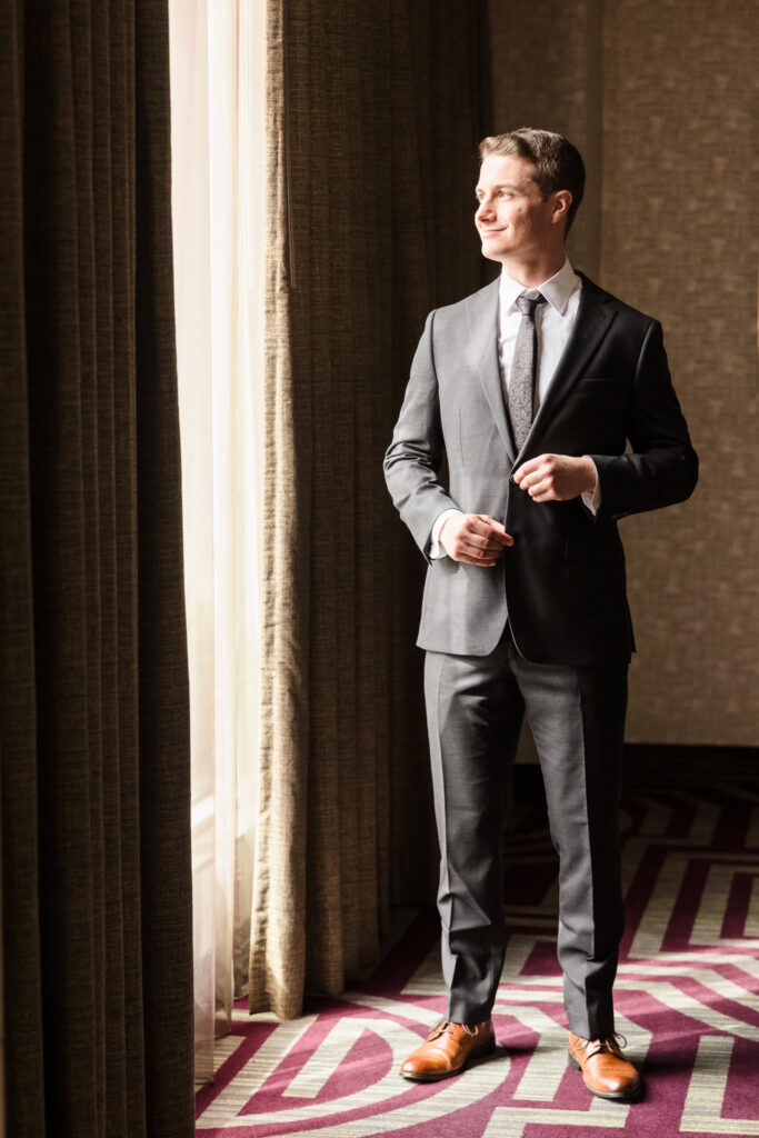 Groom standing by a window adjusting his suit jacket before the wedding ceremony.