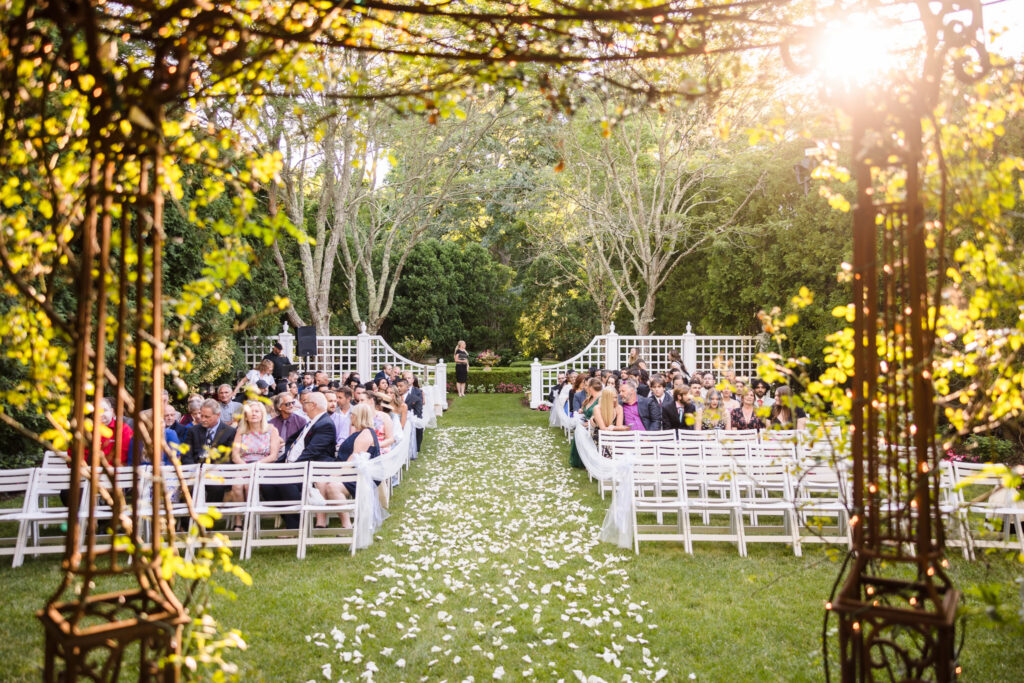 Outdoor garden wedding ceremony setup with white chairs, flower petals, and guests arriving before the ceremony.