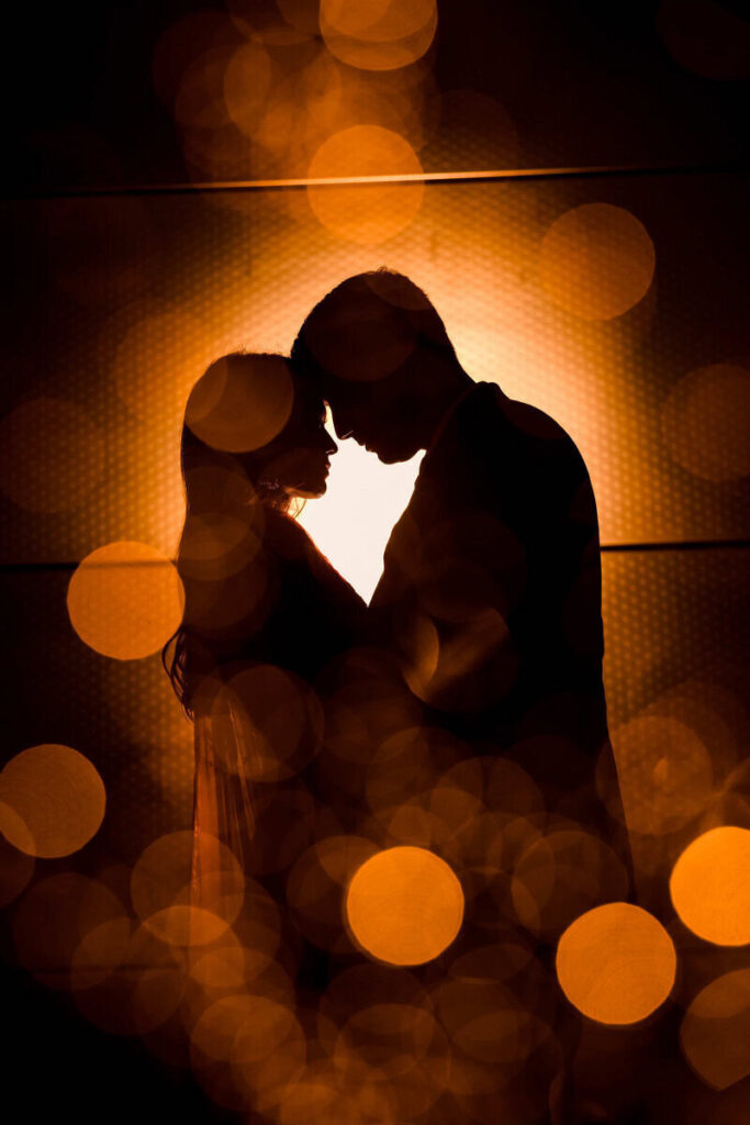 Romantic silhouette portrait of a couple embracing during their wedding reception with warm glowing lights.