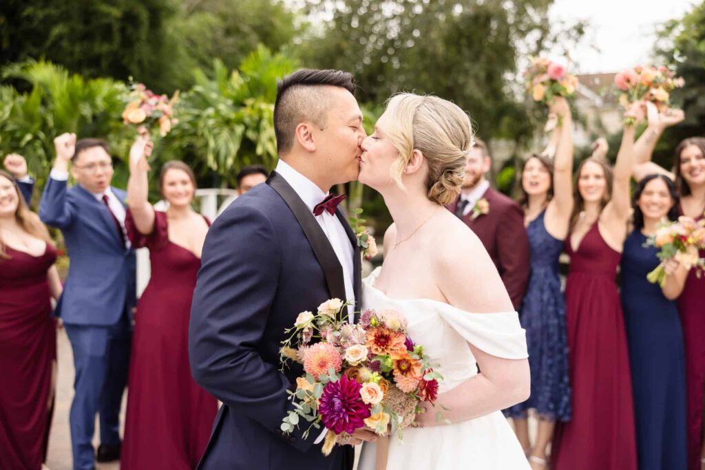 Newlyweds sharing a kiss surrounded by their cheering wedding party holding colorful bouquets.