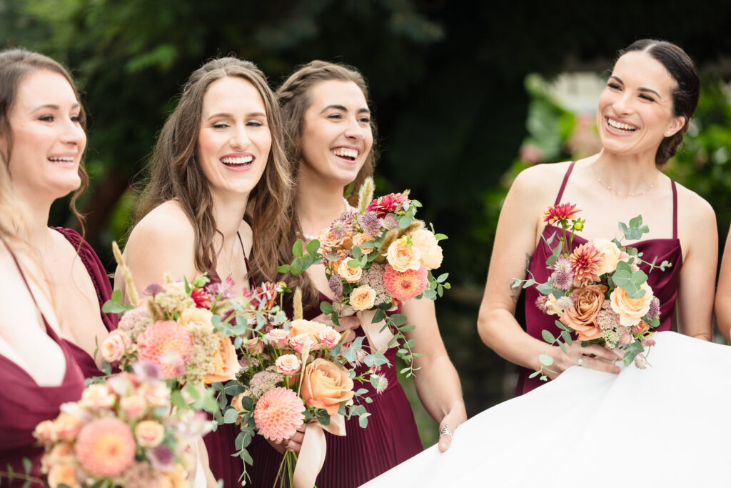 Bridesmaids laughing together while holding colorful floral bouquets during an outdoor wedding.