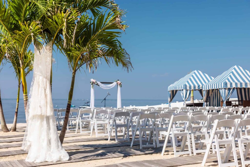 Beachfront wedding ceremony setup with white chairs, palm trees, and ocean views on a sunny day.