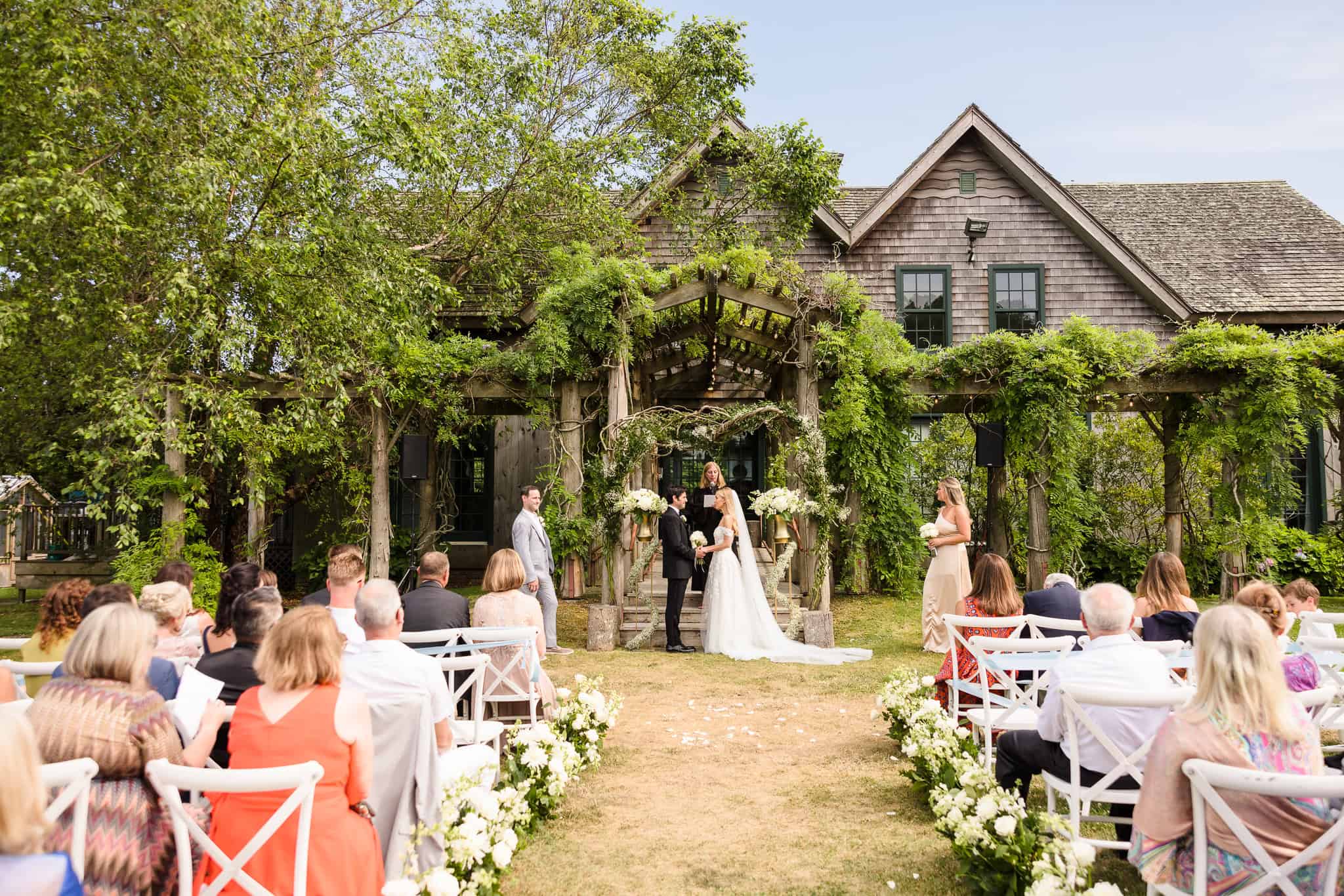 Outdoor wedding ceremony photographed at a refined Long Island venue with guests gathered beneath a vine-covered arbor.