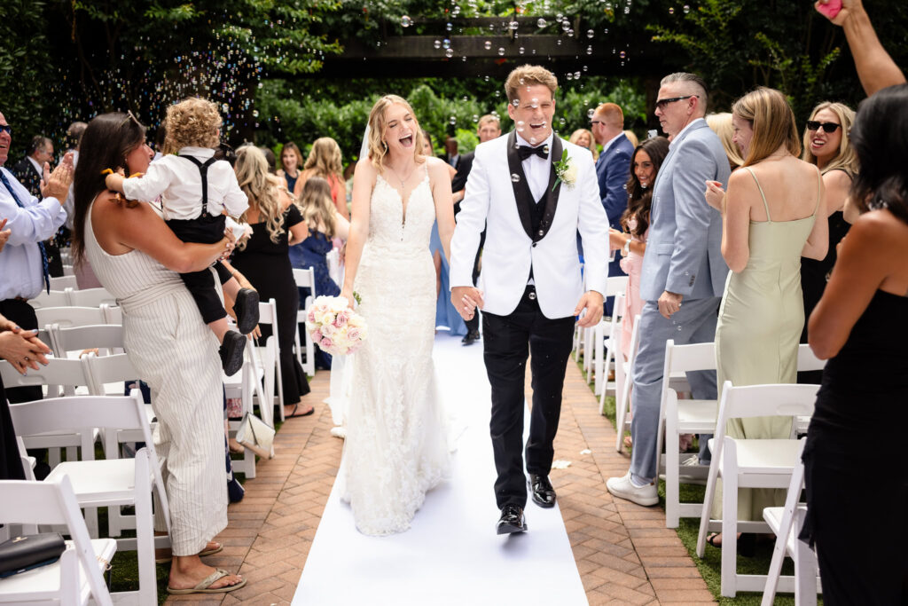 Newly married couple walking down the aisle during their wedding recessional while guests blow bubbles and celebrate.