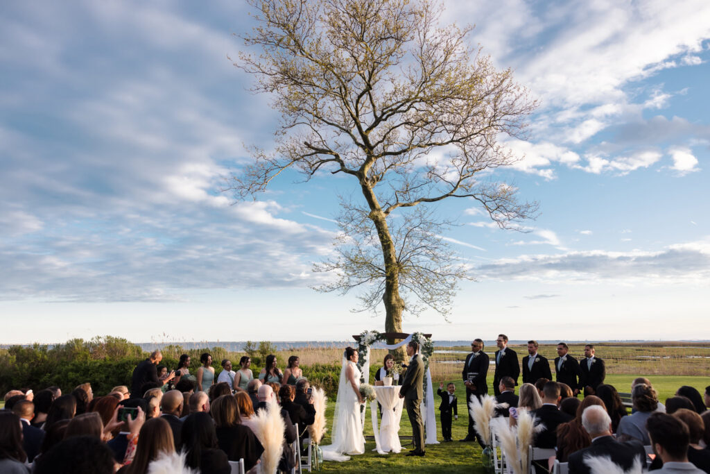 Outdoor waterfront wedding ceremony on Long Island with guests gathered beneath a large tree overlooking the water.
