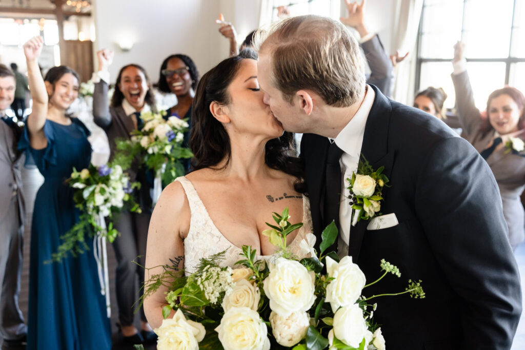 Newlyweds sharing their first kiss surrounded by cheering wedding party members.