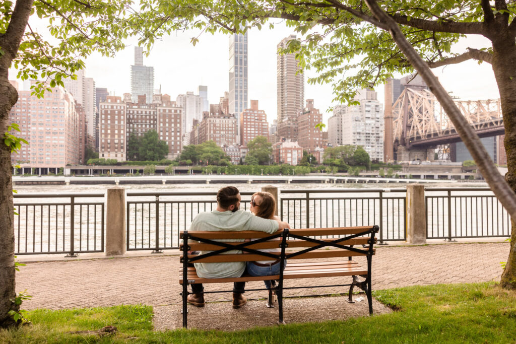 Couple sitting together along the East River during a Roosevelt Island engagement session with the Manhattan skyline in the background.