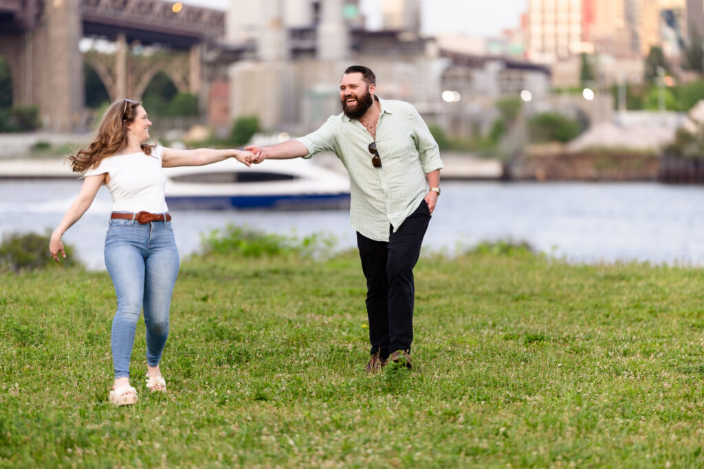 Heidi and Tom running hand in hand through a grassy waterfront park near Roosevelt Island at golden hour.