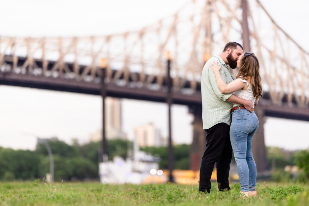 Couple kissing beneath the Queensboro Bridge during their Roosevelt Island engagement session along the East River.