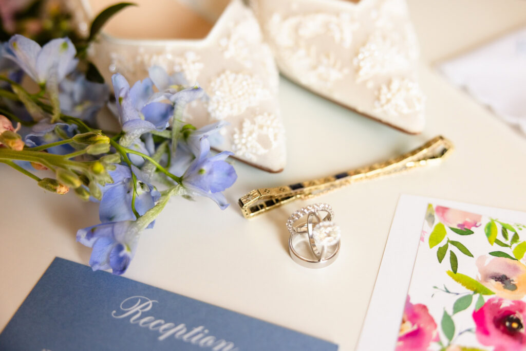 Wedding details including the bride's lace shoes, wedding rings, invitation suite, and blue florals photographed before Ashley and Kyle's Heritage Club wedding.