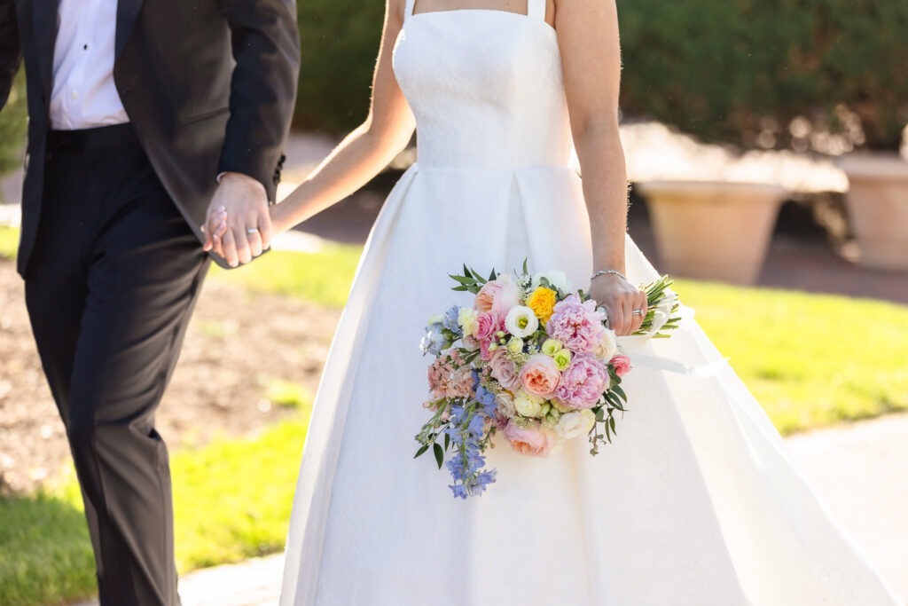 Bride and groom walk hand in hand outdoors at The Heritage Club at Bethpage, with the bride holding a pastel bouquet of pink, blue, yellow, and white flowers against her elegant wedding gown.