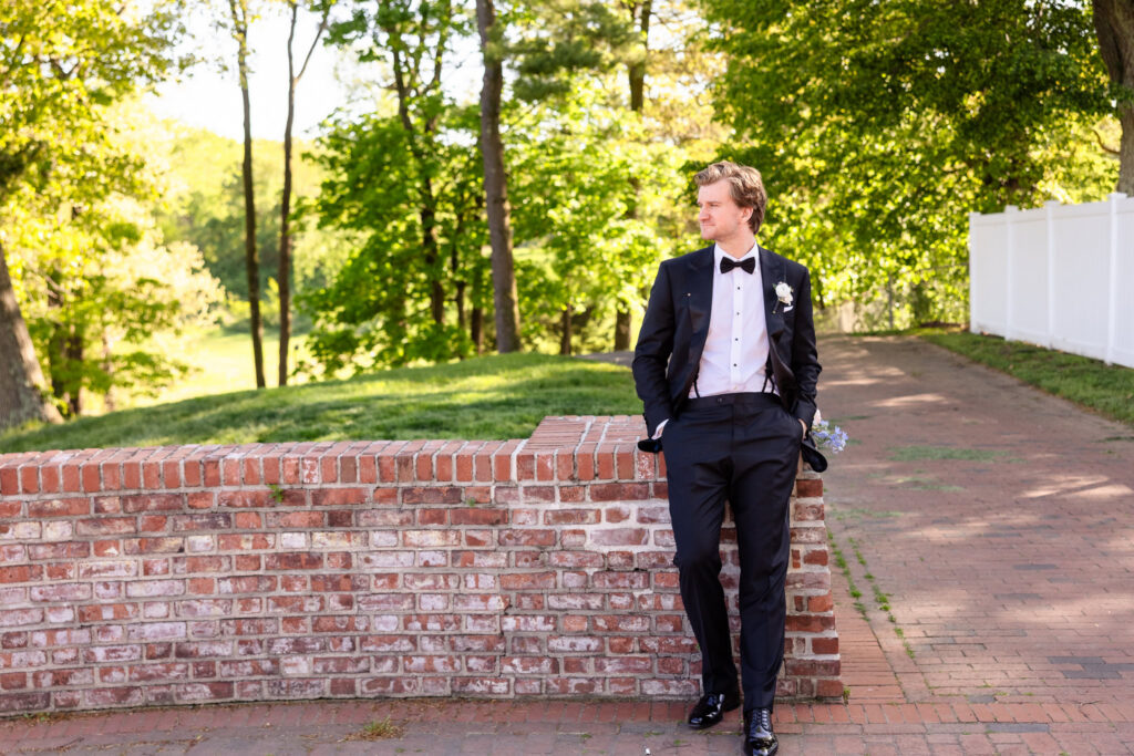 Portrait of the groom in a black tuxedo leaning against a brick wall at The Heritage Club at Bethpage, surrounded by lush green trees and soft afternoon light.