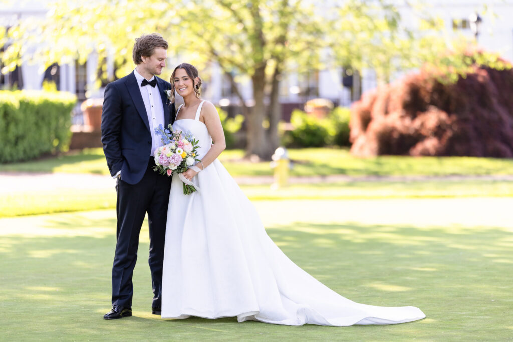 Bride and groom pose together on the golf course at The Heritage Club at Bethpage, with the bride’s long train flowing behind her and a colorful spring bouquet in hand.