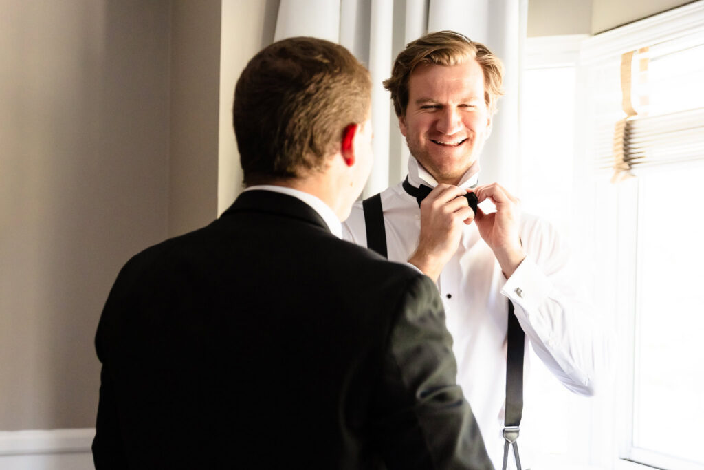 Kyle adjusts his bow tie with the help of a groomsman while getting ready at the Garden City Hotel before the wedding.