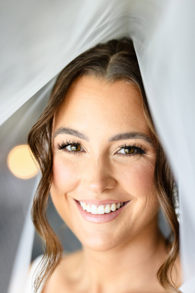 Close-up bridal portrait of Ashley smiling beneath her veil at The Heritage Club at Bethpage, highlighting her wedding makeup, soft curls, and elegant expression.