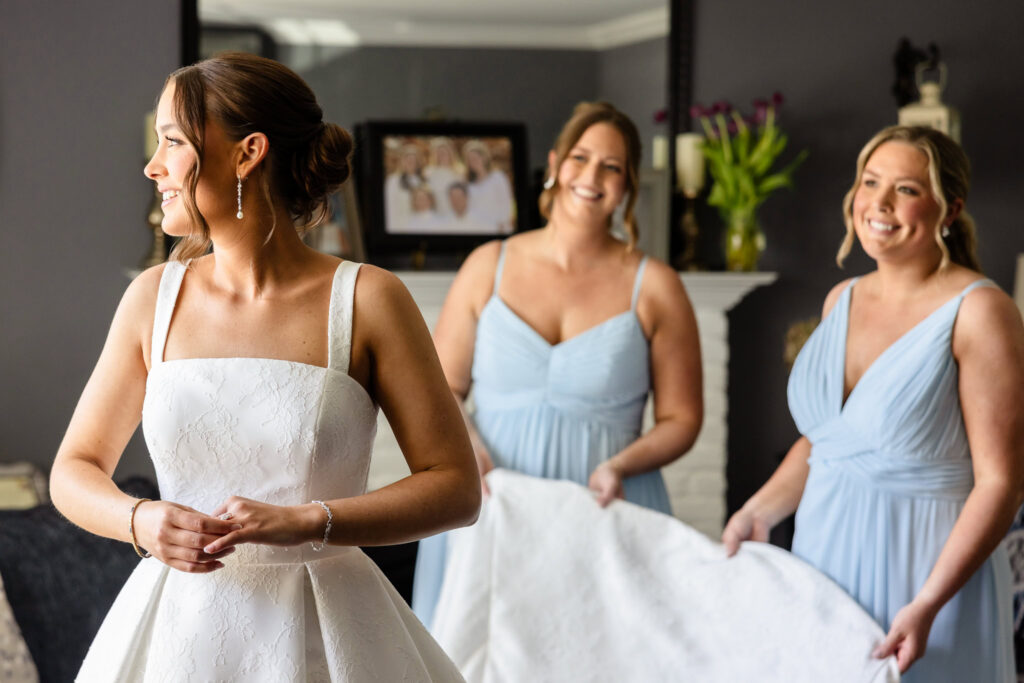 Ashley stands in her wedding dress while her bridesmaids arrange her gown during bridal preparations before her Heritage Club wedding.