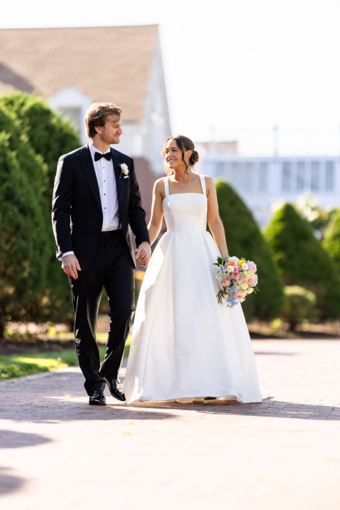 Ashley and Kyle walk hand in hand outdoors at The Heritage Club at Bethpage, smiling at each other during their wedding portraits.