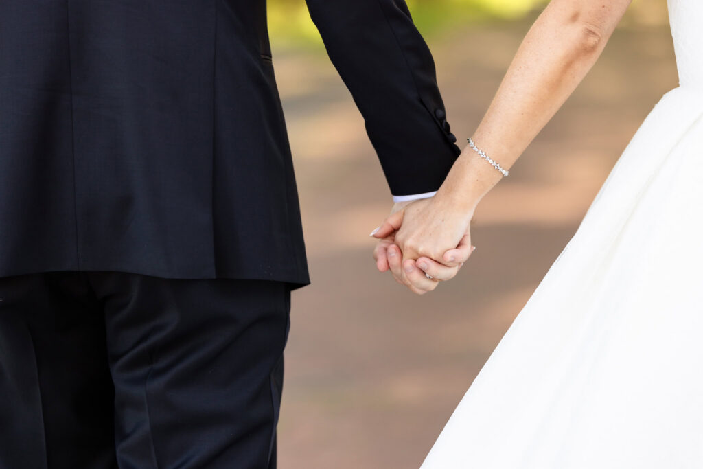 Close-up detail of Ashley and Kyle holding hands during portraits at The Heritage Club at Bethpage, highlighting the bride’s wedding band and bracelet against their elegant wedding attire.