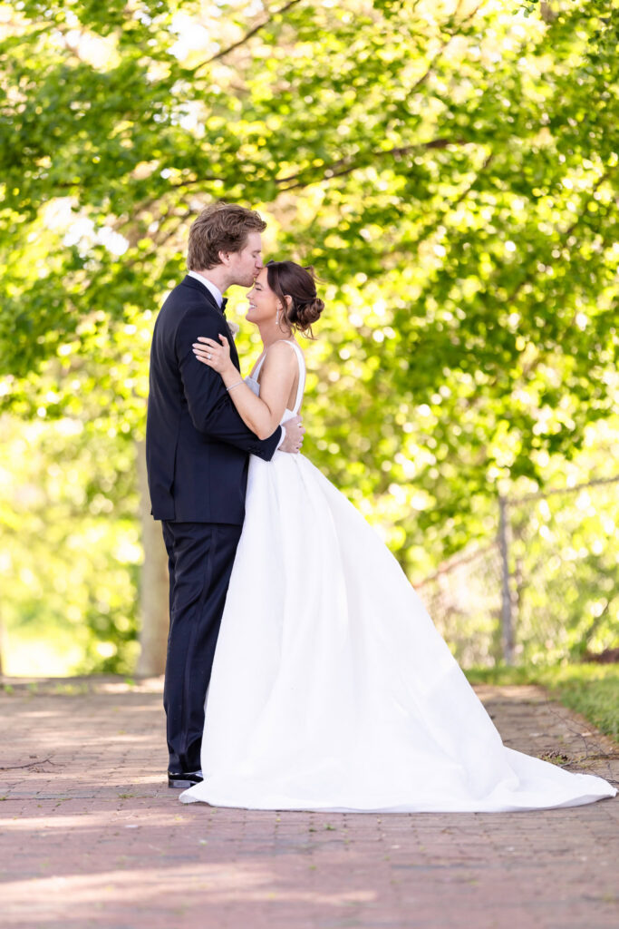 Kyle kisses Ashley on the forehead during a wedding portrait at The Heritage Club at Bethpage with lush green trees in the background.