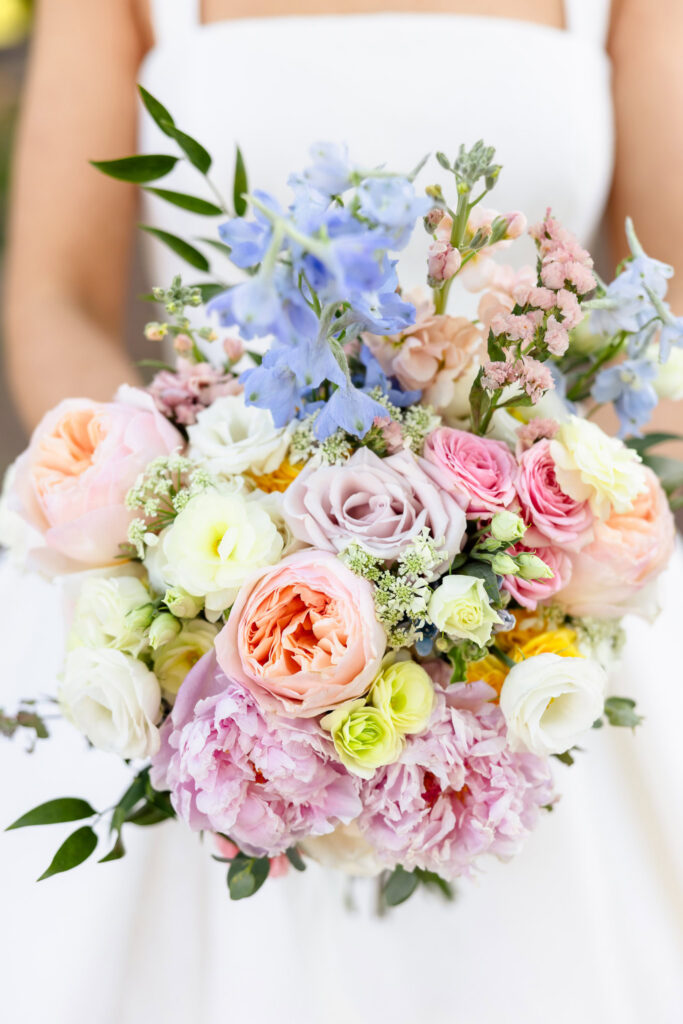 Close-up detail of Ashley’s wedding bouquet featuring blush roses, pink peonies, white blooms, and light blue delphinium flowers, photographed against her wedding gown at The Heritage Club at Bethpage.