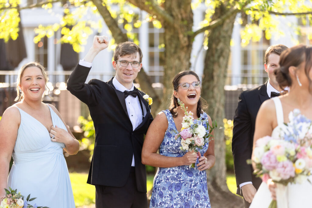 Members of the wedding party react with excitement and laughter as Ashley and Kyle share a kiss during portraits at The Heritage Club at Bethpage, surrounded by colorful spring bouquets and golden afternoon light.

