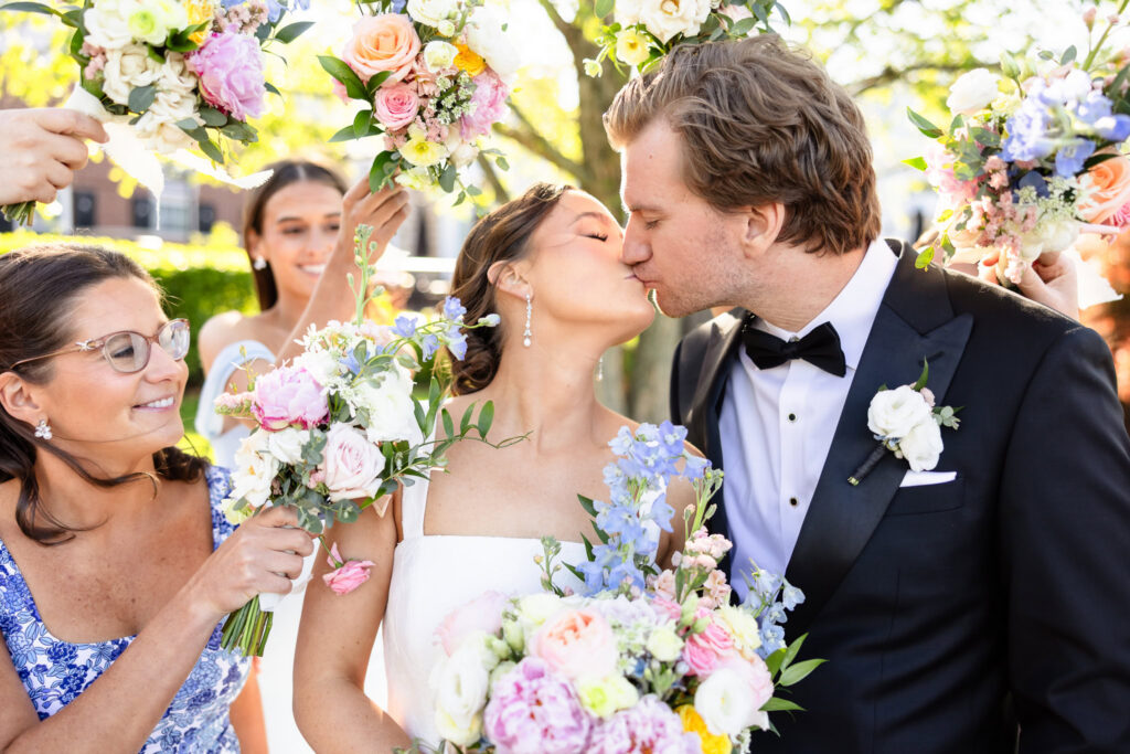 Ashley and Kyle share a kiss surrounded by bridesmaids and family members holding colorful floral bouquets at The Heritage Club at Bethpage.