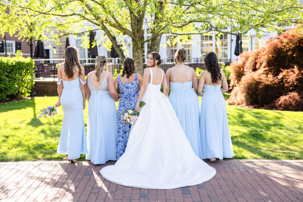 Ashley stands with her bridesmaids outdoors at The Heritage Club at Bethpage, looking back over her shoulder as the group faces away from the camera beneath a blooming tree, showcasing their coordinated light blue dresses and spring bouquets.