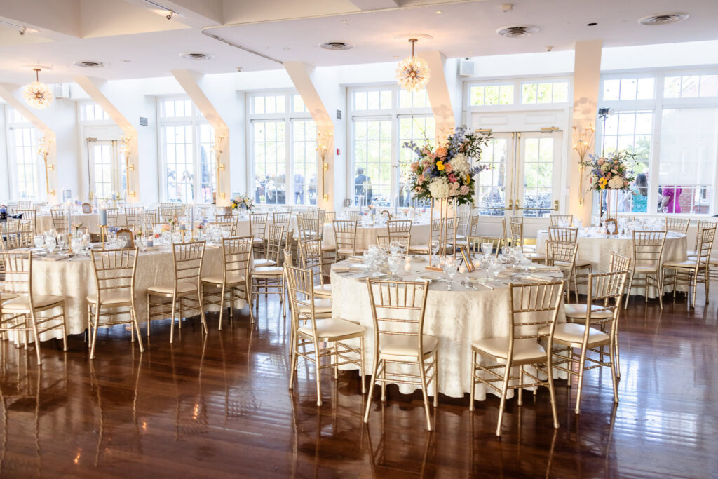 Reception ballroom at The Heritage Club at Bethpage featuring elegant floral centerpieces, gold chiavari chairs, and floor-to-ceiling windows.