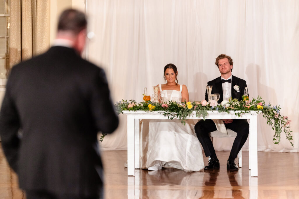 Ashley and Kyle sit at their sweetheart table during the wedding reception at The Heritage Club at Bethpage while listening to a heartfelt speech.