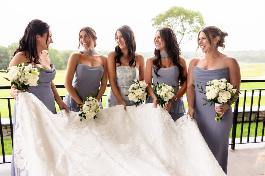 Bridesmaids smiling together while holding the bride's gown during a Huntington Crescent Club wedding.