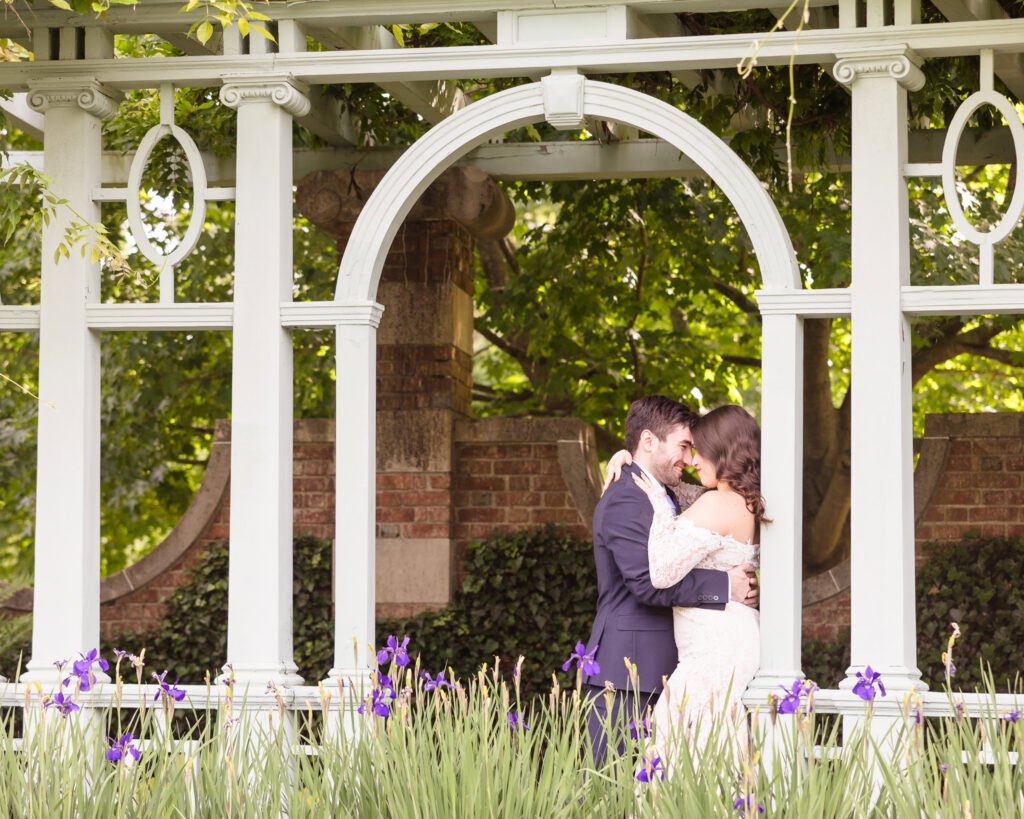 Engaged couple embracing beneath the pergola surrounded by flowers during an editorial Old Westbury Gardens engagement session.