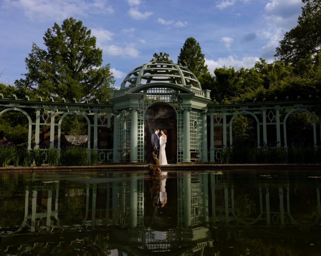 Wide portrait of a couple reflected in the pond beside the garden pavilion during an editorial Old Westbury Gardens engagement session.