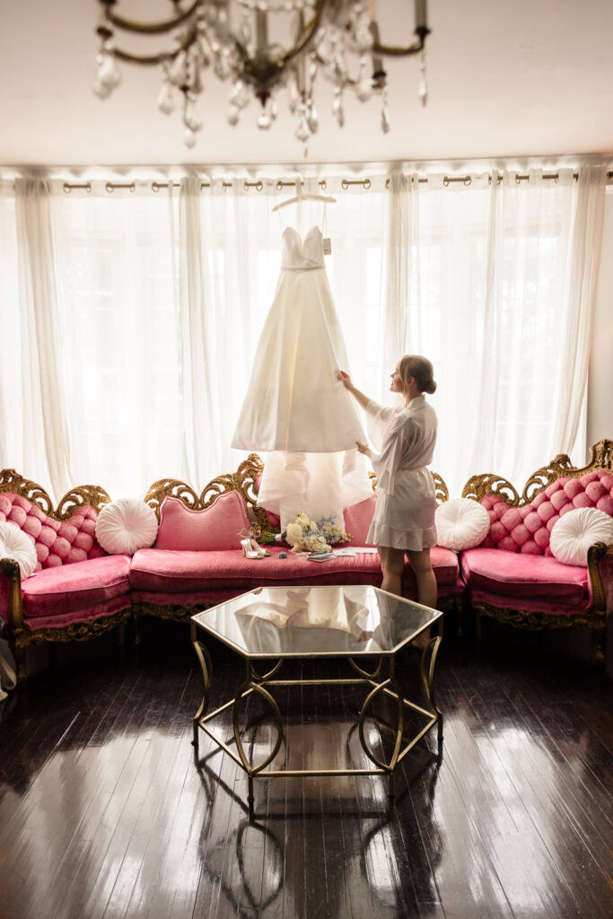Lydia admires her Suzanne Neville wedding dress hanging in front of the windows at Tarrytown House Estate before the ceremony.
