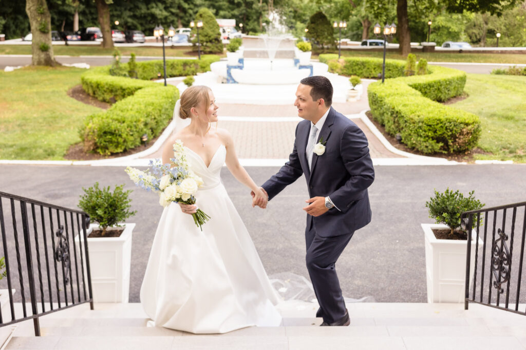 Lydia and Robbie hold hands and smile as they walk up the Briarcliff Manor staircase following their first look before the ceremony.