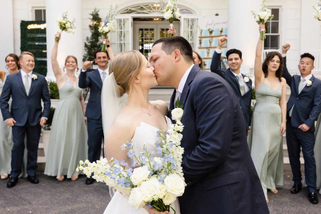 Lydia and Robbie kiss while their wedding party cheers behind them outside Briarcliff Manor after portraits.