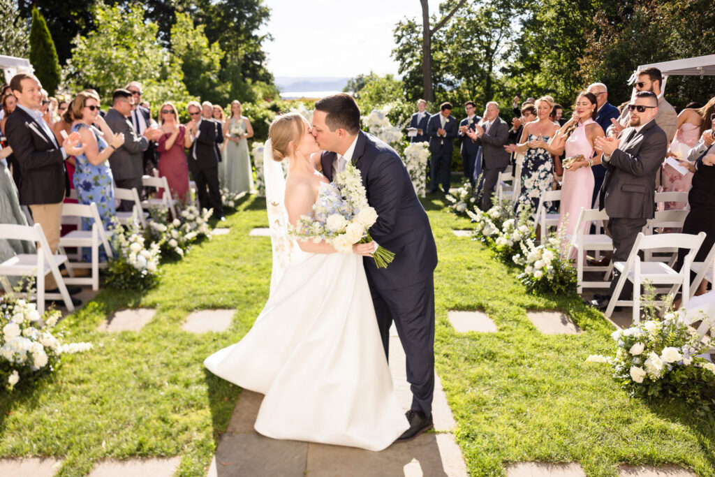 Lydia and Robbie share their first kiss as newlyweds while guests applaud during their outdoor ceremony at The Briarcliff Manor.