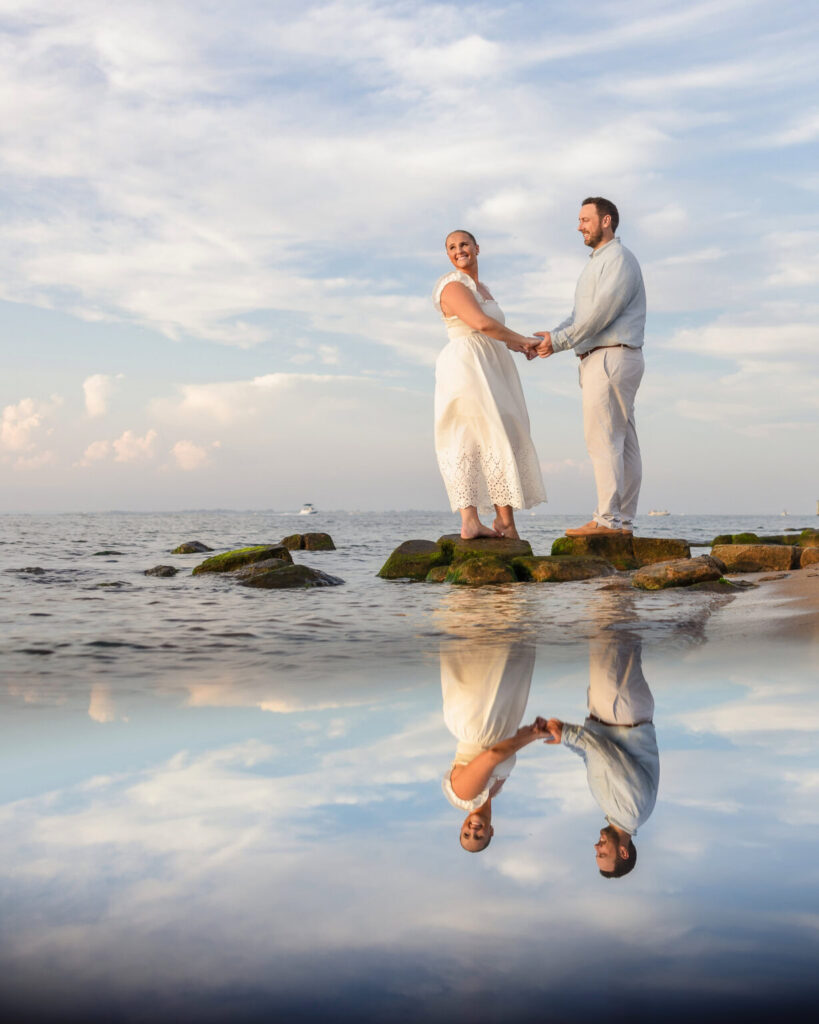 Couple holding hands on shoreline rocks overlooking the bay at Robert Moses State Park with reflections in the water.