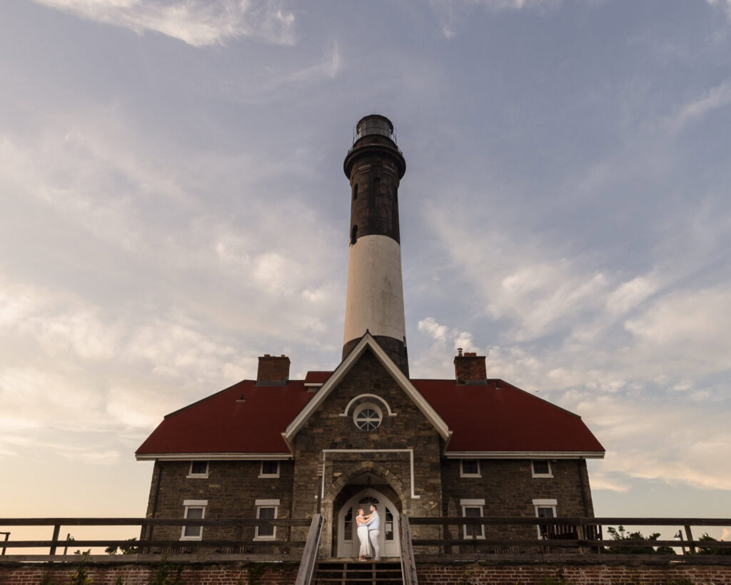 Couple embracing beneath the entrance of the historic Fire Island Lighthouse with dramatic evening skies overhead.