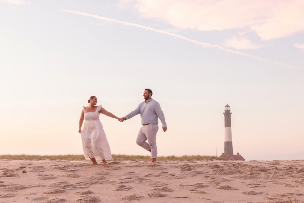 Couple walking hand in hand across the sand with the Fire Island Lighthouse in the background at sunset.
