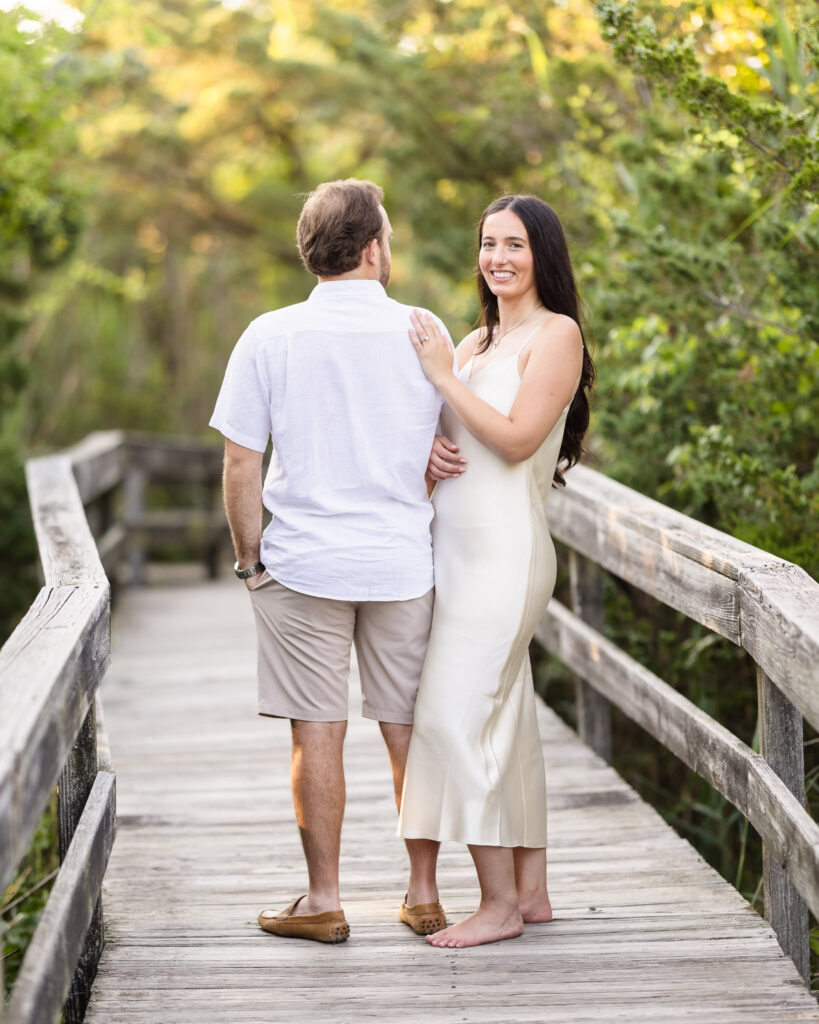 Catie smiles back at the camera while holding Zach's arm as they walk along the boardwalk at Fire Island National Seashore during their engagement session.